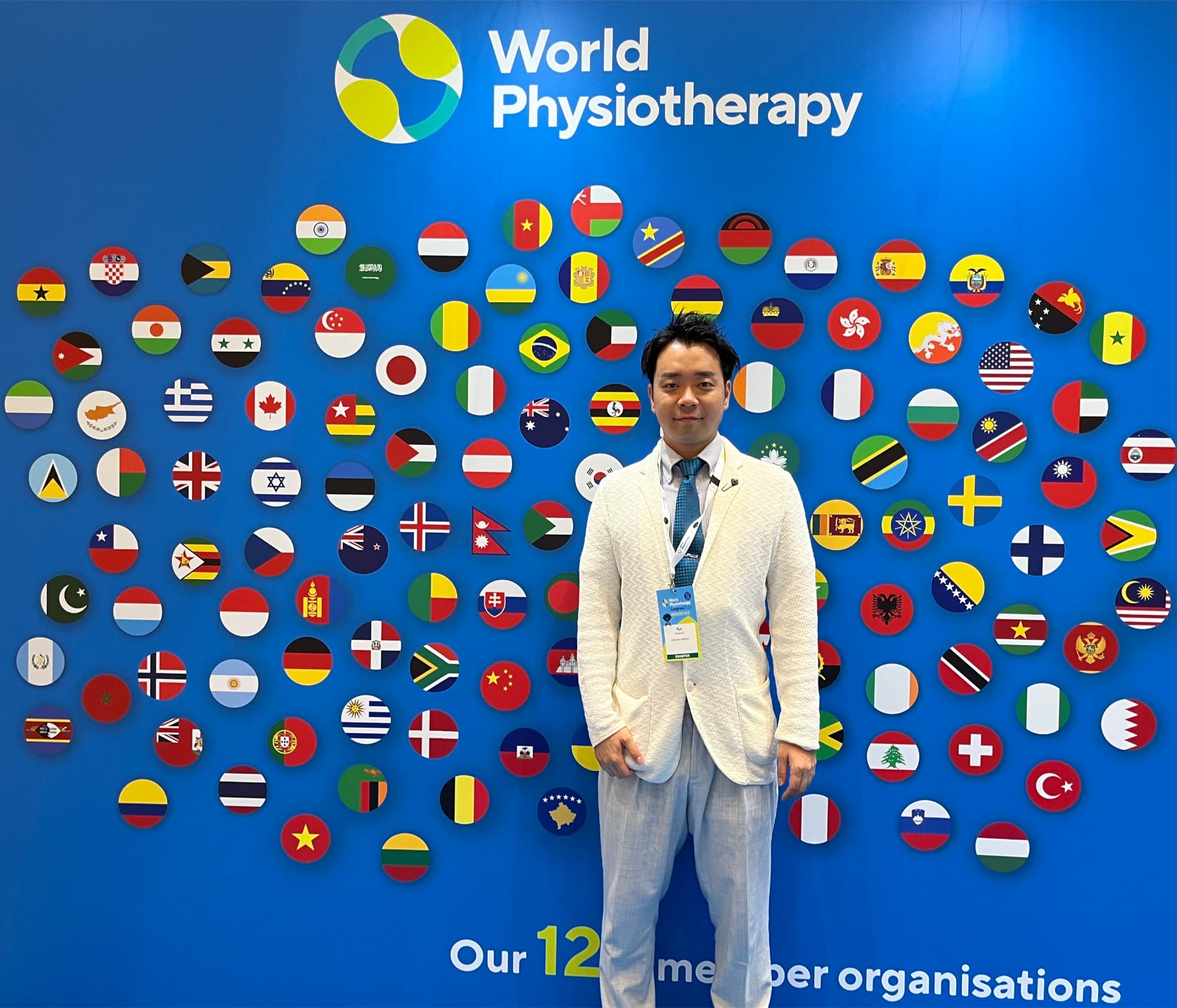 World Physiotherapyのロゴと世界の国旗が並ぶ会場フォトウォールの前に立つ藤井翔悟（出展者パス着用）